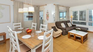 Un coin repas avec une table en bois dressée pour quatre personnes, entourée de chaises blanches. On trouve également un canapé et une petite table avec une lampe dans l’espace salon.