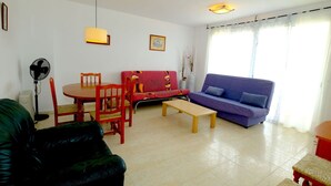 Ein Wohnzimmer mit einem roten Sofa, einem blauen Sofa, einem Holzespeisetisch mit Stühlen, einem schwarzen Ledersessel, einem kleinen Couchtisch, einem Standventilator und einem Fenster mit Vorhängen.