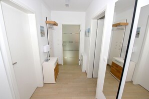 A narrow hallway with white walls, a wooden floor, and a mirror reflecting a bathroom with a sink and a towel rack.
