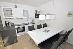 Moderni keittiö, jossa on valkoiset kaapit, sisäänrakennettu uuni sekä ruokailutila, jossa on valkoinen pöytä ja tuolit.
