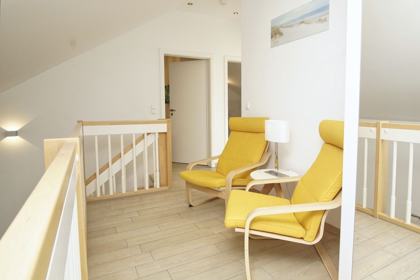 A room with a wooden staircase, two yellow chairs, a small white table with a lamp, and a painting on the wall.
