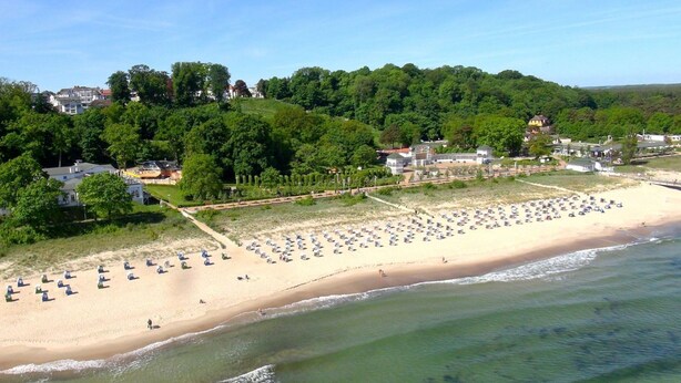 A beach with numerous umbrellas and sun loungers, a sandy shore, and a forested hill in the background. - Quiet location in Ostseebad Göhren, veranda, - Villa Möwe FeWo 03 - Villa Möwe (Göhren)