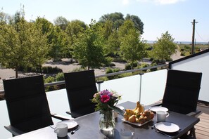 A balcony with a table set for two, featuring a vase of flowers, a fruit plate, and a cup.