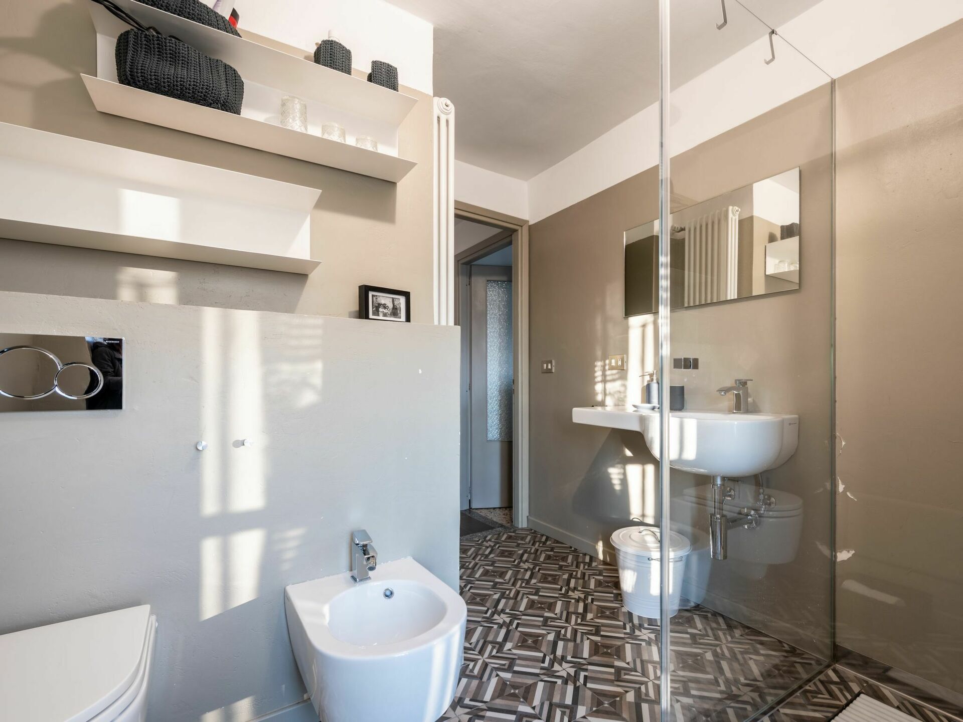 Un bagno moderno con bidet, lavandino e specchio. Sopra il lavandino ci sono delle mensole e un quadro appeso al muro.