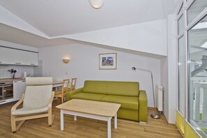 A modern living room with a green sofa, a white armchair, and a wooden coffee table. There is a dining area with a table and chairs, and a kitchen in the background. - With a view of the sea and a balcony, access to the pool and sauna - Sea View Apartment 17 - (Göhren)