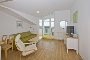 Ein modernes Wohnzimmer mit einem grünen Sofa, einem Holztisch und einem weißen Sessel. Es gibt einen kleinen Schreibtisch mit Fernseher und Computer. Der Raum verfügt über große Fenster und einen Balkon mit Blick.