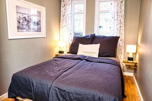Una cama bien hecha con una colcha oscura y una almohada que dice "Lieblingsplatz". Se ven dos mesitas de noche con lámparas, y un cuadro enmarcado cuelga en la pared.