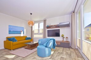 A modern living room with a yellow sofa, a blue armchair, a wooden coffee table, and a large TV.