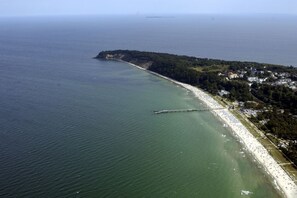 An aerial view of a coastal area with a long pier extending into the sea, a sandy beach, and a densely wooded peninsula.