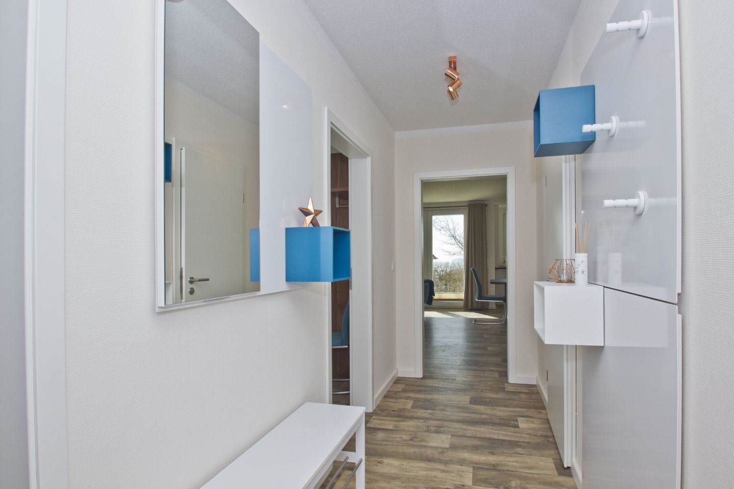 A narrow hallway with a wooden floor, white walls, and a large mirror on the left. There are blue storage boxes on the walls and a white bench at the end of the hallway.