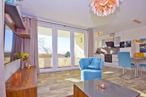 A modern apartment with a large sliding glass door leading to a balcony, a kitchen area, and a seating area with a blue armchair and a table.