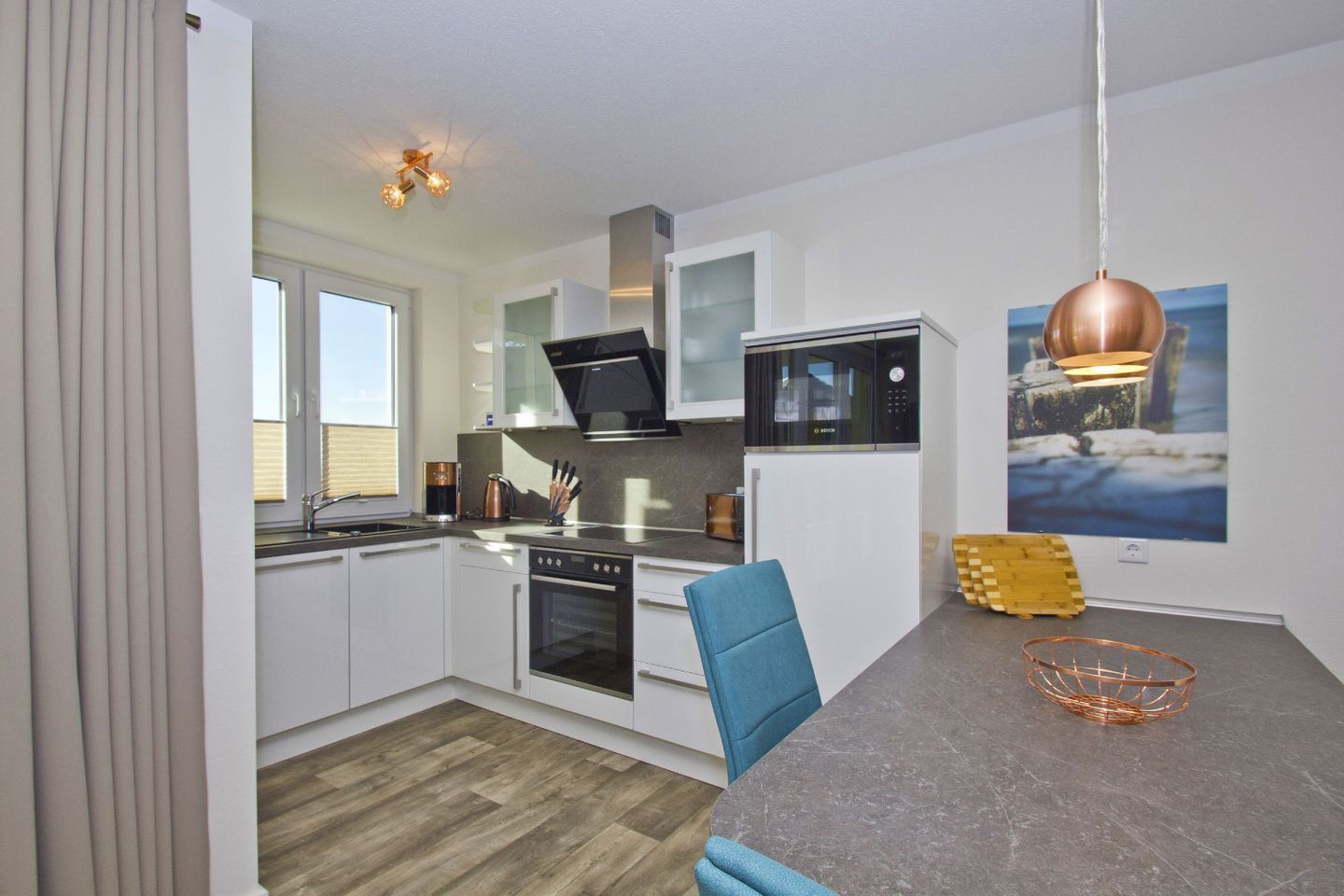 A modern kitchen with white cabinets, a Bosch oven, a microwave, and a coffee machine. There is a dining table with blue chairs and a copper-colored basket.