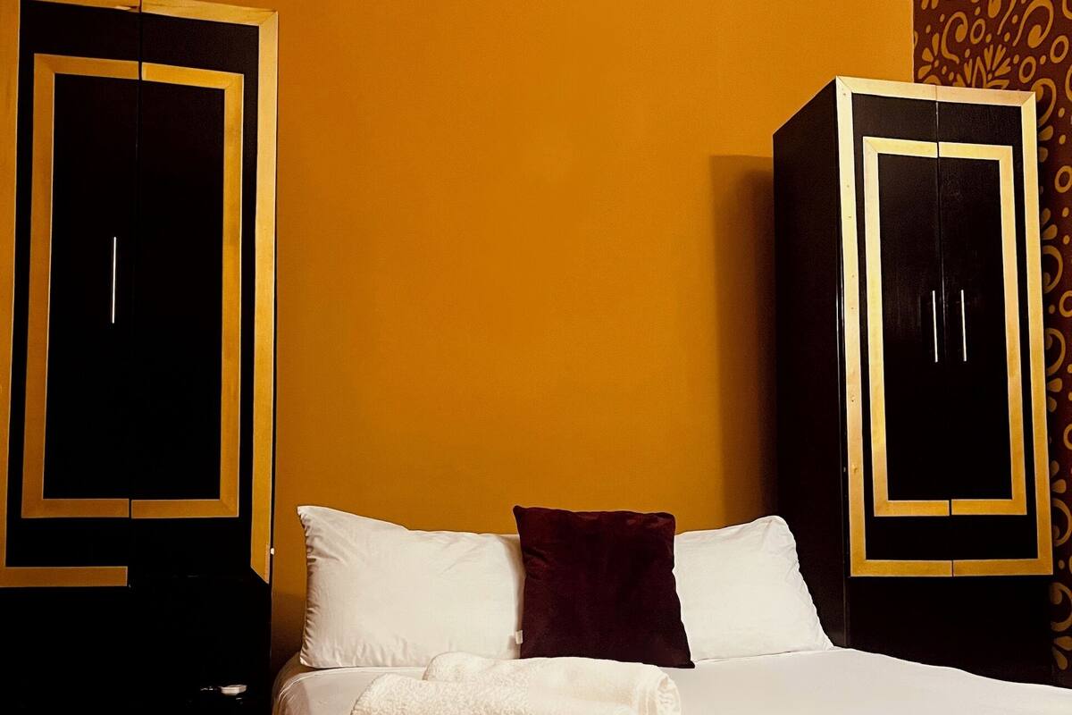 A hotel room with a bed, white pillows, a dark red pillow, and two wardrobes with gold trim.