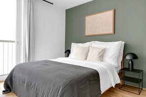 Une chambre moderne avec un grand lit, deux tables de chevet, une œuvre d’art abstraite encadrée et une vue sur l’extérieur.