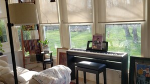 Une pièce confortable avec un piano, un bureau, une chaise, une grande fenêtre avec des stores et un ventilateur de plafond.