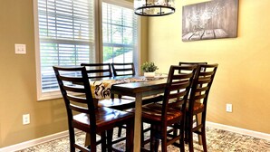 Un comedor con una mesa y sillas de madera, una lámpara colgante, una ventana con persianas y un cuadro en la pared.