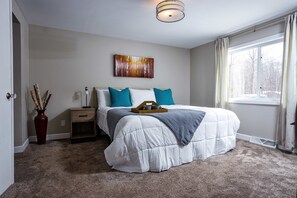 A bedroom with a large bed, a bedside table, a nightstand, a vase with decorative sticks, a painting on the wall, and a window with curtains. - The Reserve at Plum Bottom in Egg Harbor! (Egg Harbor)