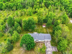 Une vue aérienne d’une maison entourée d’une forêt dense, avec une allée bien visible et une petite construction sur la droite.