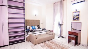 Un dormitorio con cama, mesita de noche, armario y un ventilador de pie.