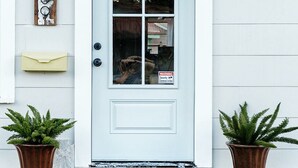 A white front door with a warning sign, two potted plants, and a mailbox on the wall.
