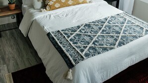 A neatly made bed with white linens, a patterned throw blanket, and a decorative mirror on the wall.