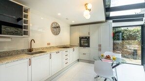 Een moderne keuken met witte kasten, een granieten aanrechtblad en een ingebouwde oven. Er is een eethoek met een glazen tafel en stoelen, en uitzicht op het landschap door een groot raam.