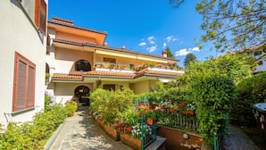 A courtyard with a stone pathway leading to a building with balconies, surrounded by lush greenery and flowering plants. - Appartamento Gaya 2BR with Terrace, Stresa, Italy (Stresa)
