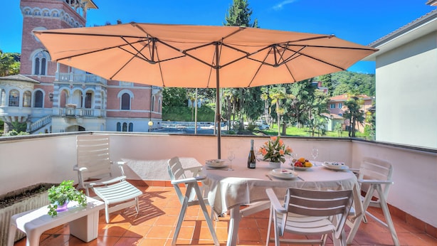 A terrace with a table, chairs, and an umbrella, overlooking a historic building and greenery. - Appartamento Gaya 2BR with Terrace, Stresa, Italy (Stresa)