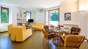 A living room with a yellow sofa, wicker chairs, a dining table with chairs, a TV, and a window with a view of greenery. - Appartamento Gaya 2BR with Terrace, Stresa, Italy (Stresa)