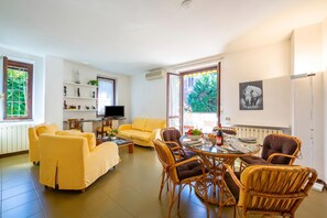 A living room with a yellow sofa, wicker chairs, a dining table with chairs, a TV, and a window with a view of greenery.
