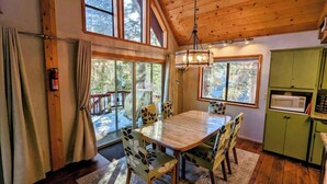 Un comedor con mesa y sillas de madera, una lámpara colgante y vistas al exterior a través de amplios ventanales.
