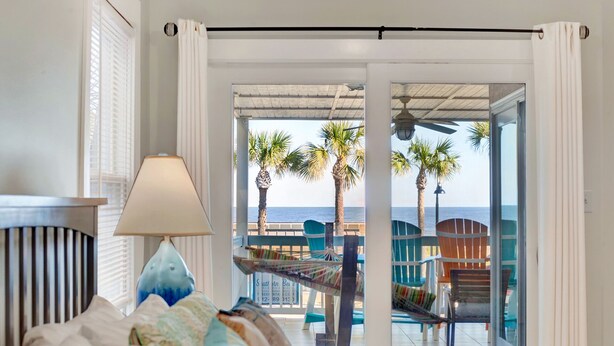 A room with a bed, a lamp, and a view of the beach with palm trees and a chair. - Gorgeous Ocean Views, Heated Pool Access (Tybee Island)