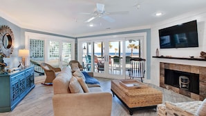 A spacious living room with a large sofa, a television, a fireplace, and a view of the beach through the glass doors. - Gorgeous Ocean Views, Heated Pool Access (Tybee Island)