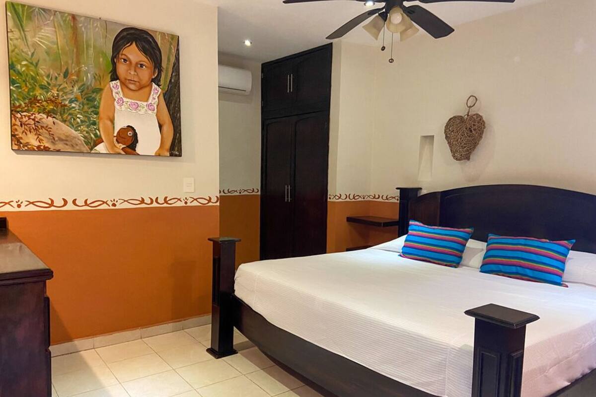 A bedroom with a large bed, a ceiling fan, a painting on the wall, and a decorative heart hanging on the wall.