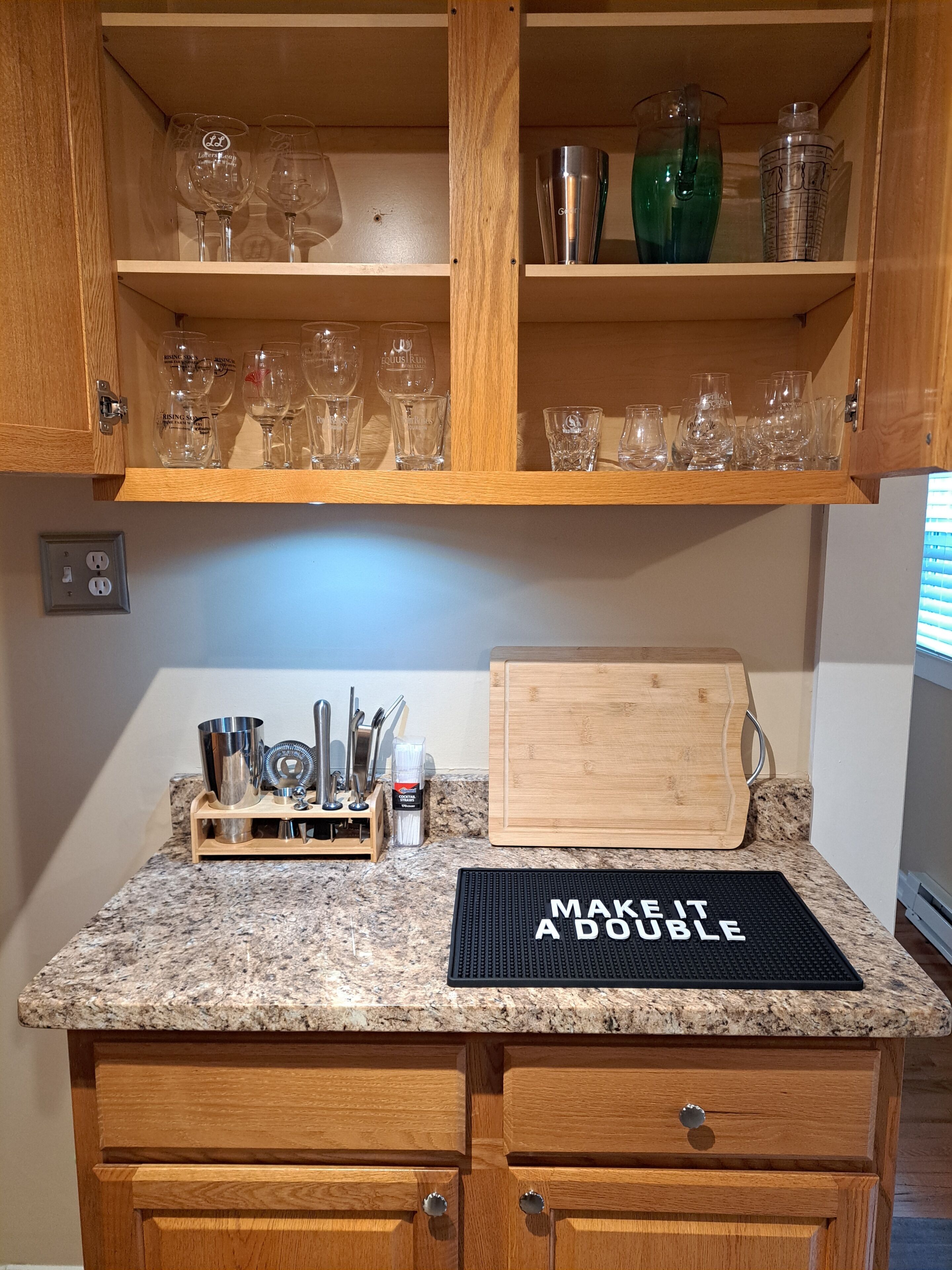 A kitchen with wooden cabinets, a granite countertop, a cutting board, and a mat with text.