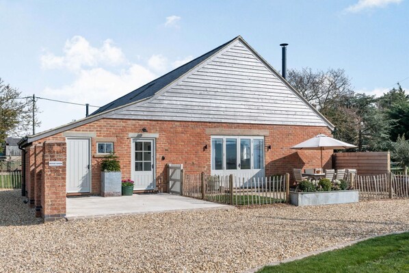 A brick building with a white door, windows, and a gravel driveway. - Dog friendly holiday home with superb garden and rural location - Ridge Barn (Honington, near Shipston-on-Stour)