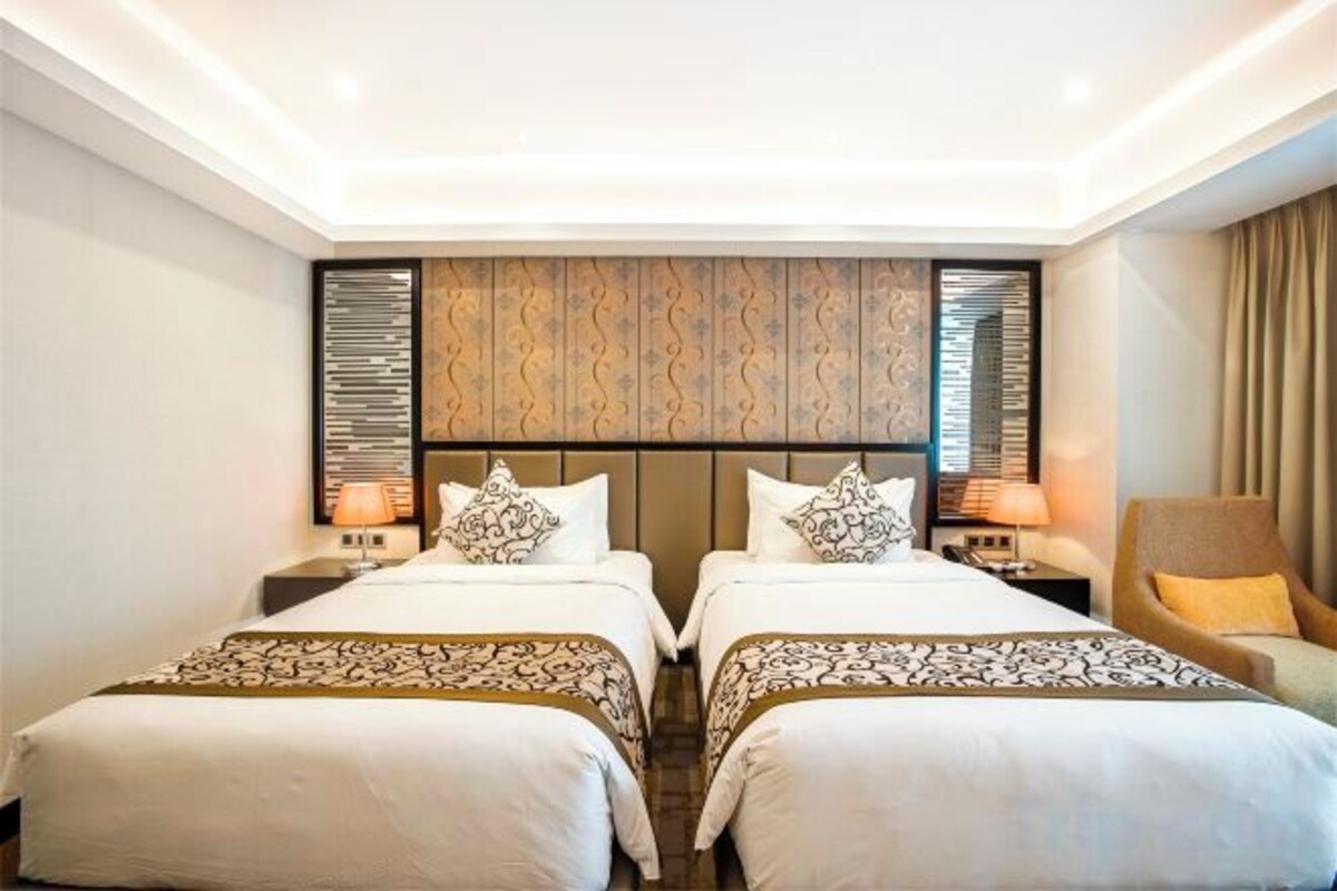 A hotel room with two beds, a headboard with a patterned design, bedside tables with lamps, and a chair.