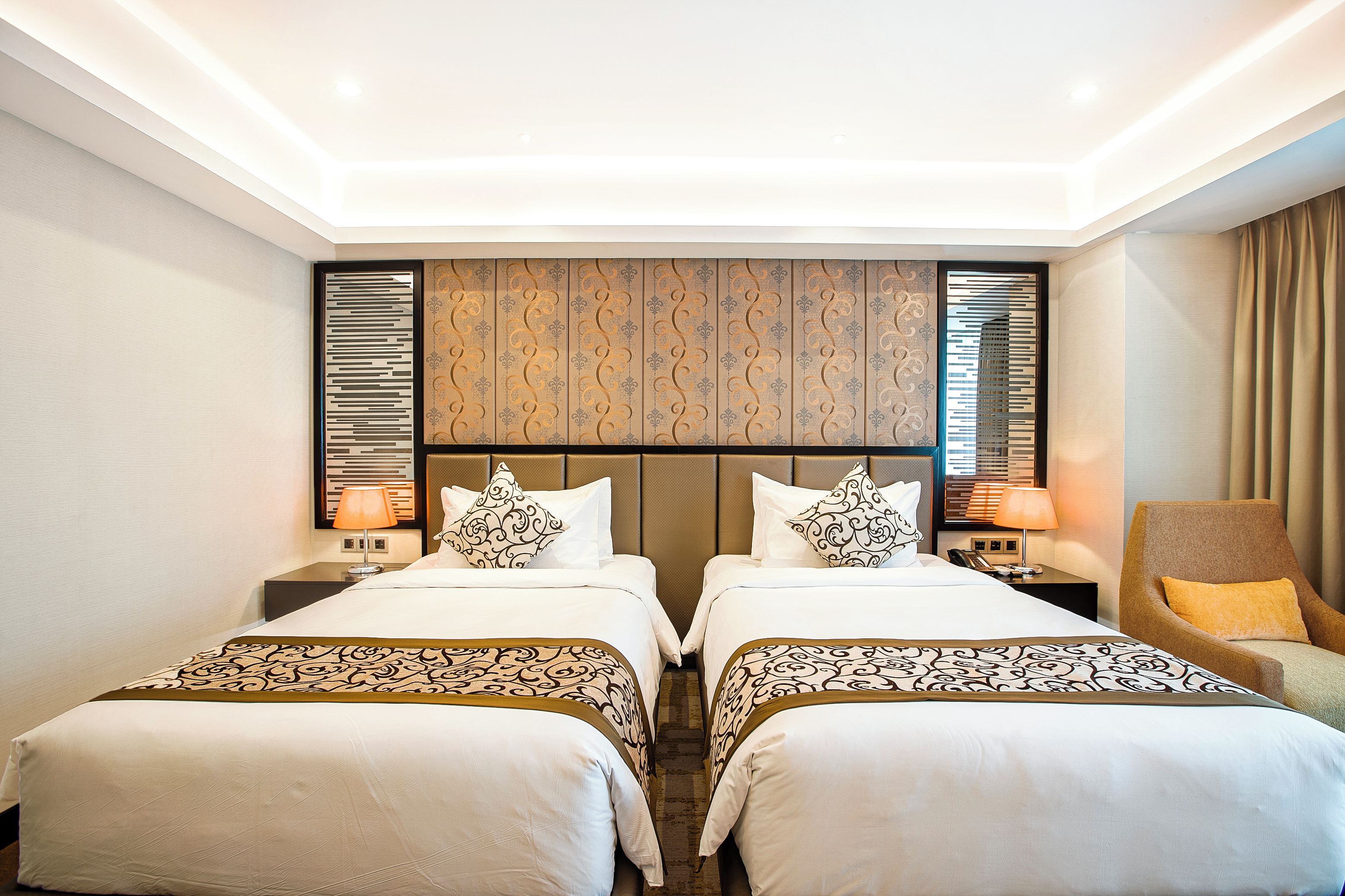 Habitación de hotel con dos camas, cabecero con panel decorativo, lámparas de noche, una silla con cojín y una cortina.