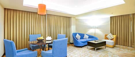 A hotel room with a modern design, featuring a round dining table with chairs, a blue sofa, a yellow armchair, and a patterned carpet.