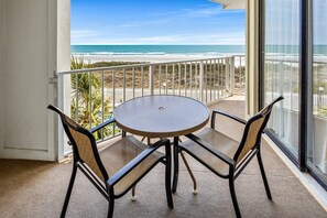 A balcony with a round table and two chairs, overlooking a beach and ocean.