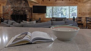 A cozy cabin interior with a stone fireplace, a large window, a sofa, a table with a bowl, and an open book.