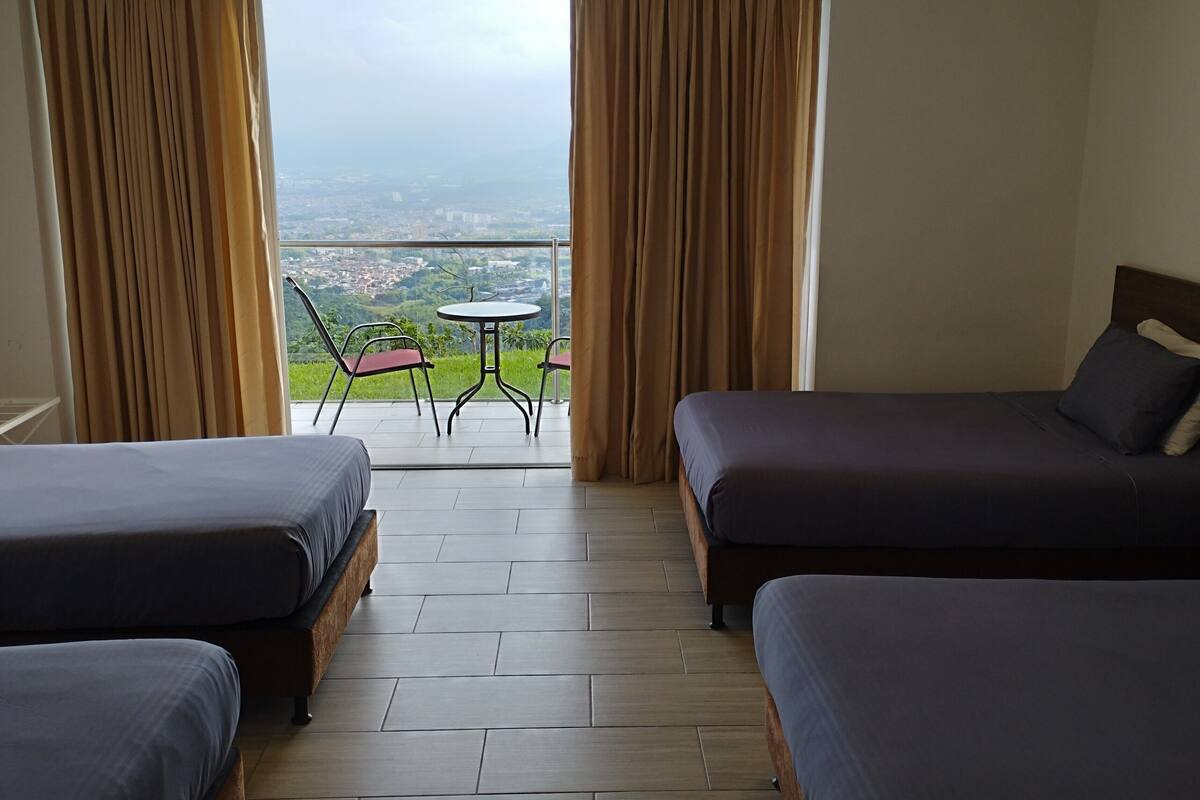 A hotel room with two beds, a small table, and chairs. The room has a view of a cityscape through large windows.