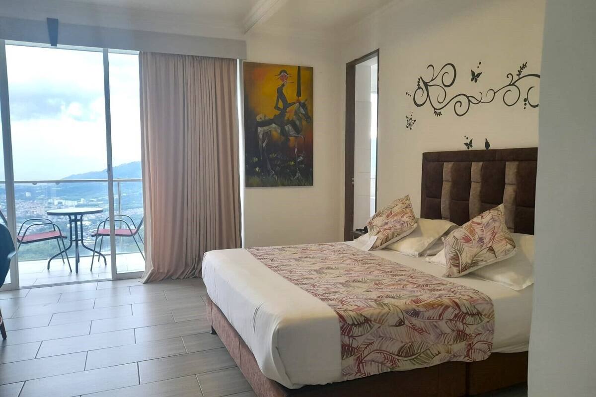 A hotel room with a large bed, a view of the city through a balcony, and a painting on the wall.