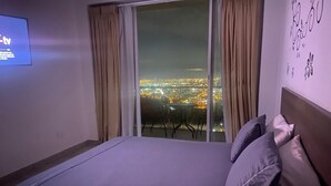 Habitación de hotel con cama, televisor en la pared y vista a la ciudad a través de amplios ventanales.