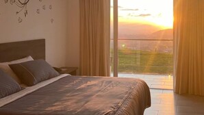 Un dormitorio con una cama grande, una mesita de noche y un adorno en la pared. Hay una puerta corrediza de vidrio que da a un balcón con vista al atardecer.