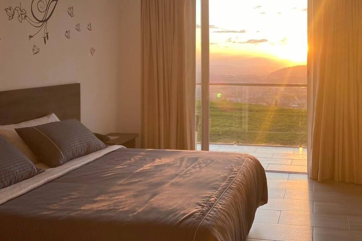 A bedroom with a large bed, a nightstand, and a wall decoration. There is a sliding glass door leading to a balcony with a view of the sunset.