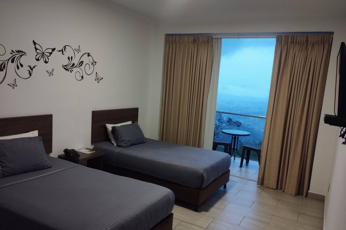 A hotel room with two beds, a small table, and a view of the sea through large windows.