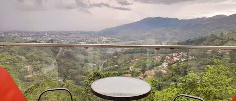 A balcony with two red chairs and a small round table, overlooking a cityscape and green hills.