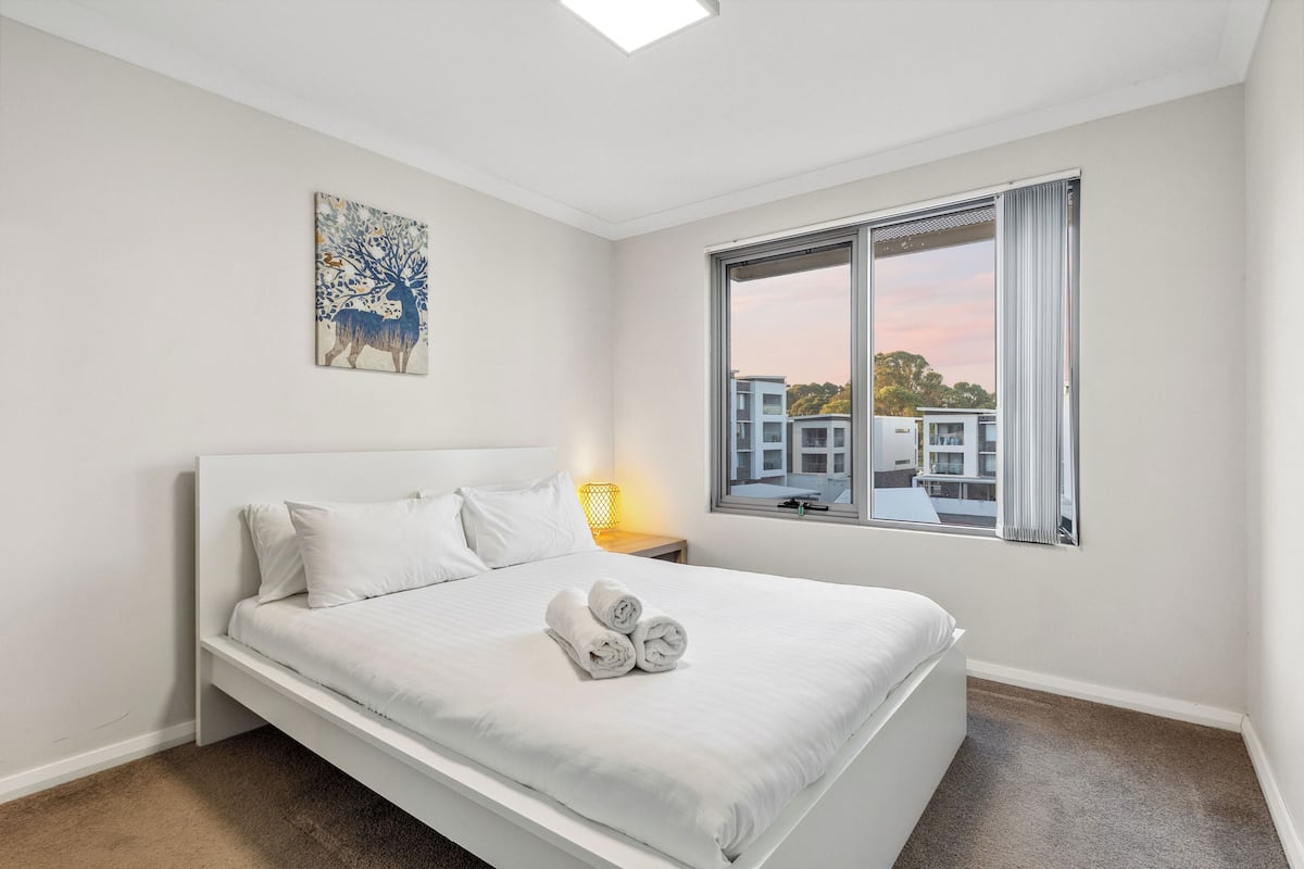 Ein modernes Schlafzimmer mit einem großen Bett, einem Fenster mit Blick auf Gebäude und einem Bild an der Wand.