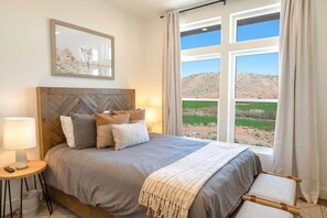 Un dormitorio con una cama grande, cabecera, mesita de noche con lámpara y vista a un paisaje a través de ventanas amplias.